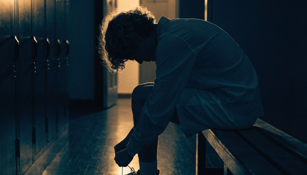 An athlete lacing a shoe on a bench in a dark locker room, seen from behind